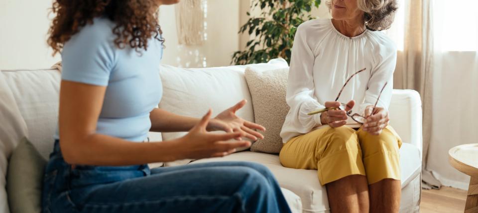 Two women sit on a couch having a conversation. One gestures expressively, while the other listens, holding glasses