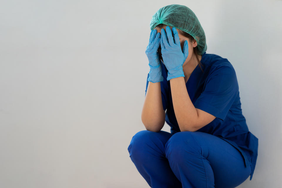 Healthcare worker in scrubs and gloves, sitting and covering face with hands, appears stressed or overwhelmed