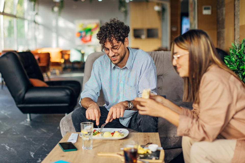 Two people enjoying a meal together at a cozy indoor café, sitting on a sofa. One is cutting food while the other holds a wrap