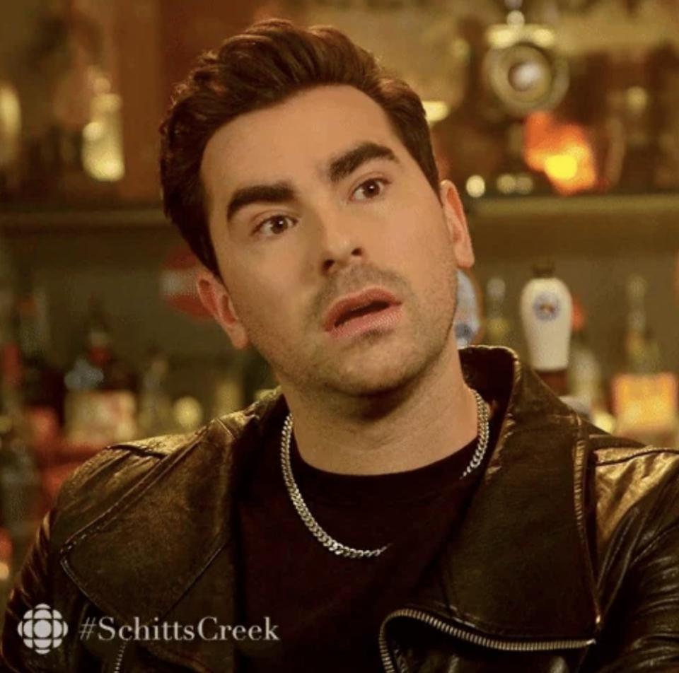 Person with short hair in a bar setting, wearing a leather jacket and chain necklace, looking surprised. #SchittsCreek logo in corner