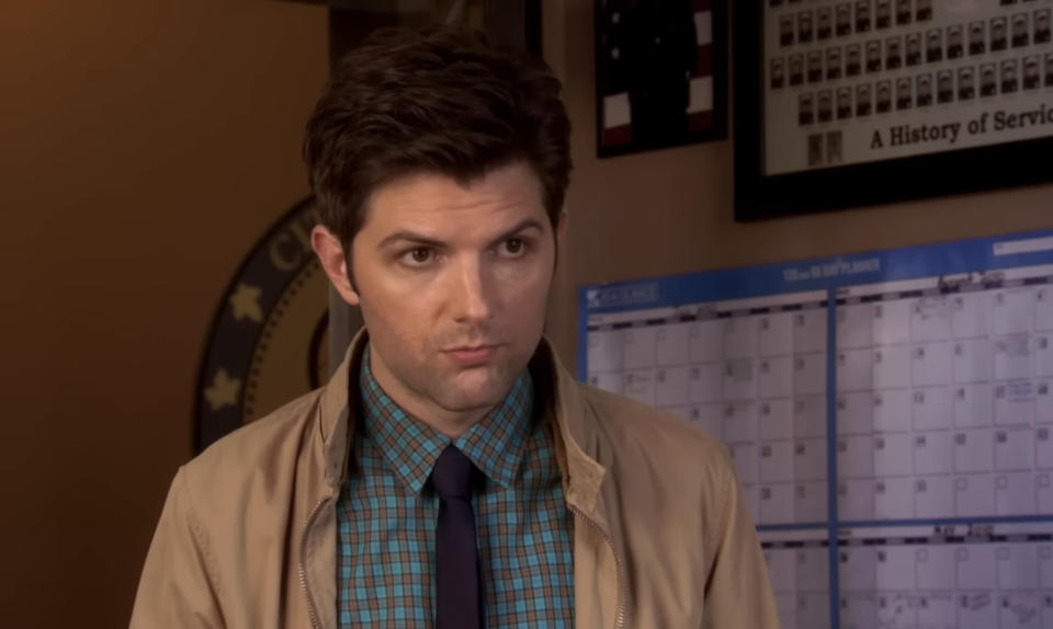 A man in a checkered shirt and tie stands indoors, looking focused. Behind him, there is a calendar on the wall