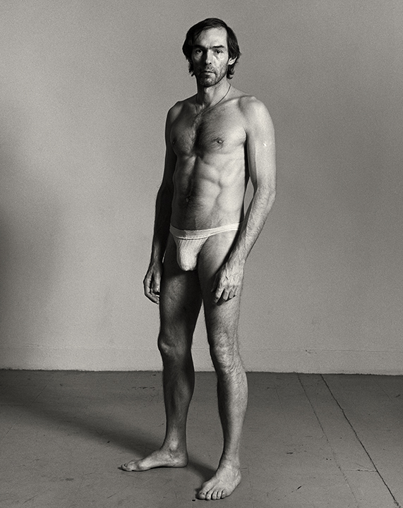 A man stands barefoot in a studio space wearing only a small white thong, his expression calm as he looks directly at the camera in a frontal self-portrait.