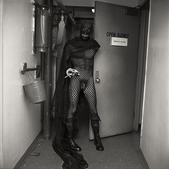 A person in a fishnet bodysuit, black boots, gloves and a full-face mask stands in a hallway holding a cigarette, partially draped in a long cape, next to a door marked “Open Slowly.”