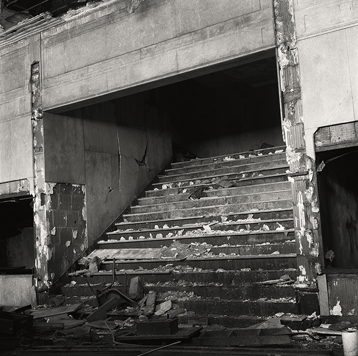A dilapidated staircase inside an abandoned building is covered in broken debris, flaking paint and rubble, evoking urban decay.