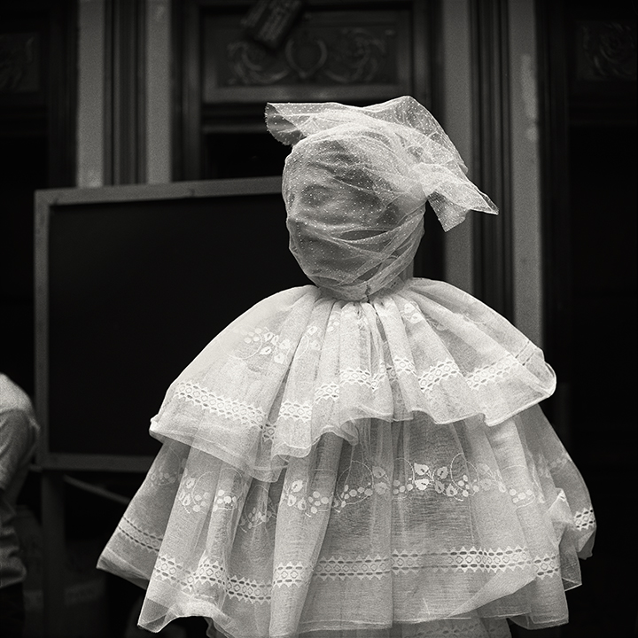 A person stands wrapped in layers of lace and sheer fabric with their face completely obscured, appearing as a costumed figure in a backstage setting.