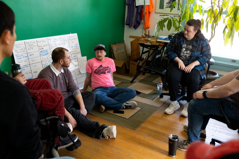 People sit in chairs and on the floor in a room with a green wall and a plant next to a window.