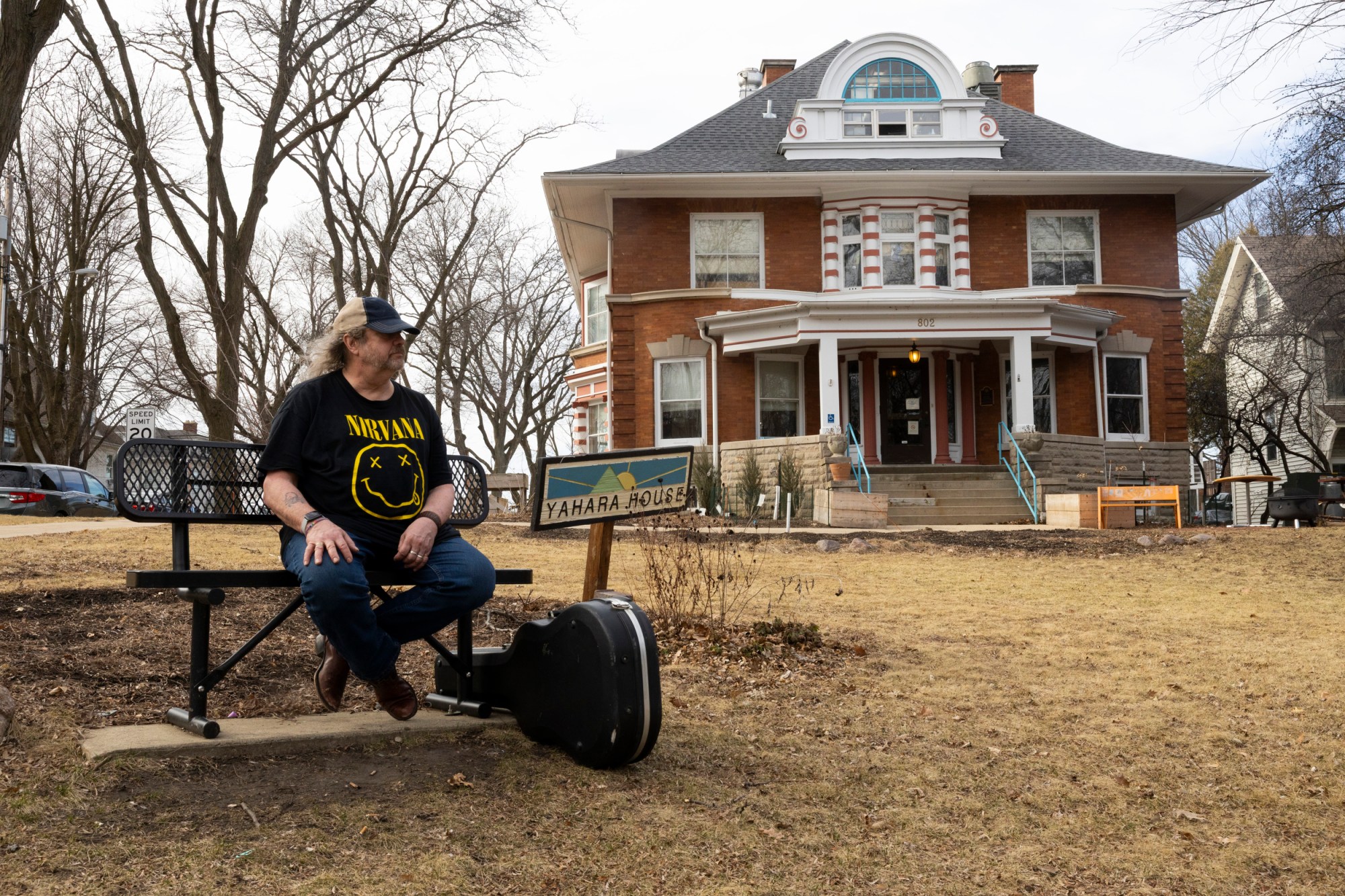 Man in Nirvana shirt sits on bench in front of house with guitar case on ground next to him.