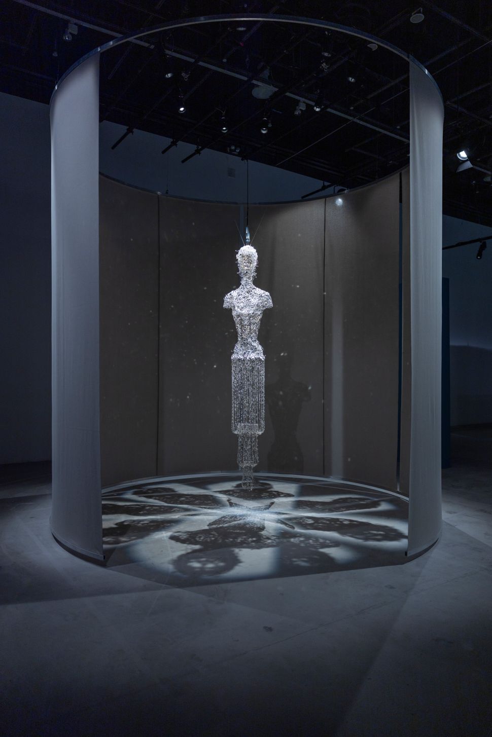 A suspended humanoid figure composed of glistening, crystalline strands hangs within a circular curtained enclosure, casting a mandala-like shadow pattern on the floor beneath it.