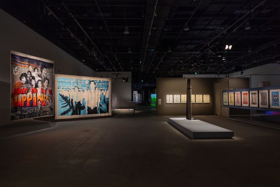 Central hanging sculpture made of colorful tangled textiles and yarns illuminated dramatically against a gray wall, casting soft, organic shadows that spread across the gallery floor.