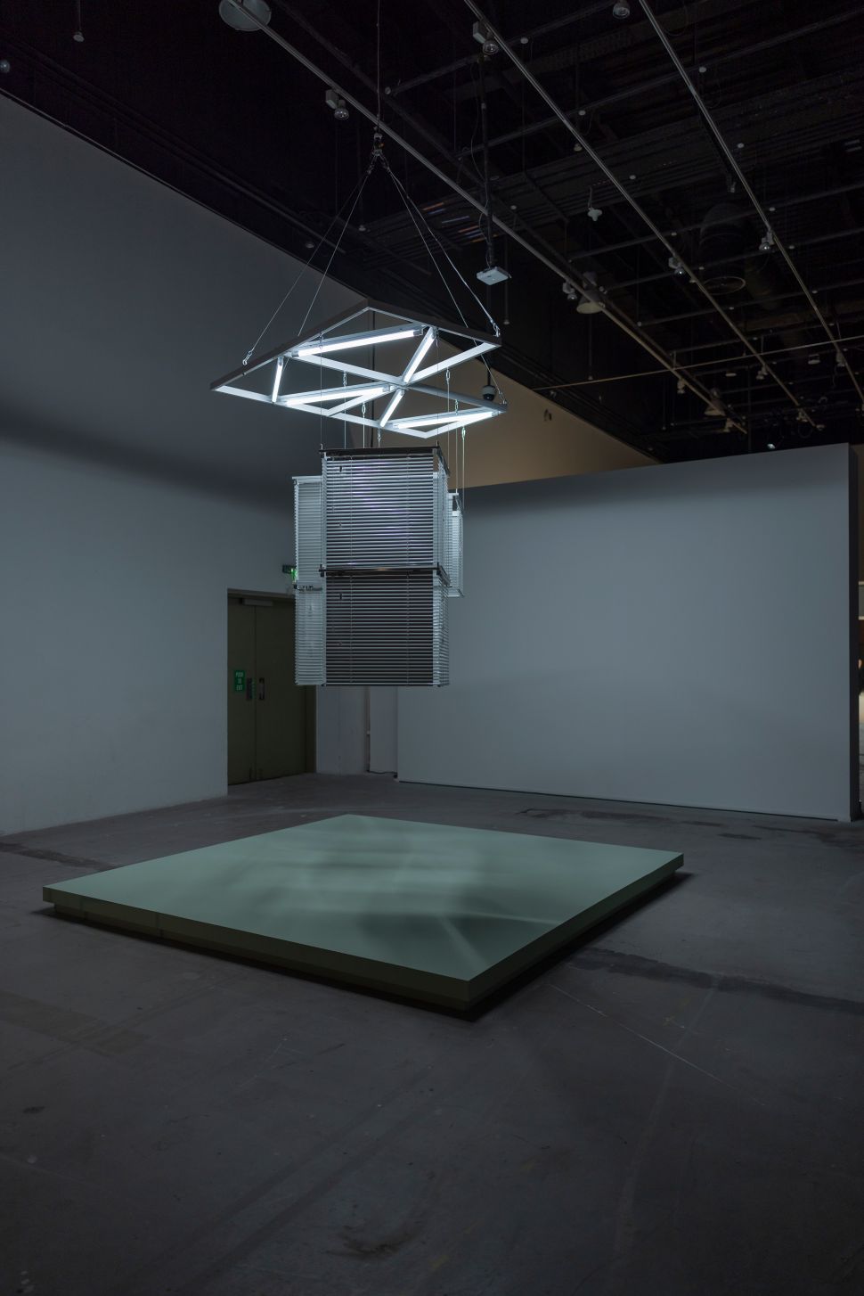Minimalist installation featuring a suspended geometric frame with fluorescent lights and metallic blinds, casting faint, rippling shadows onto a low square platform below.