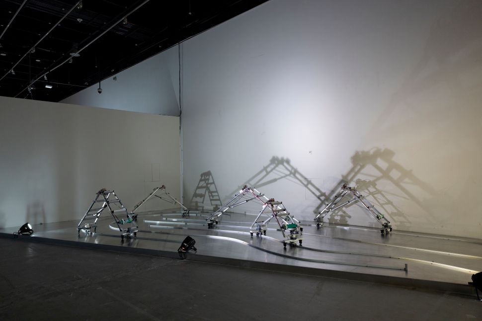 A kinetic sculpture installation featuring a group of robotic, multi-jointed metal structures moving along curved tracks on a raised platform. The metallic forms resemble insect-like walkers or skeletal ladders, casting large geometric shadows on the white gallery wall behind them under focused stage lighting.