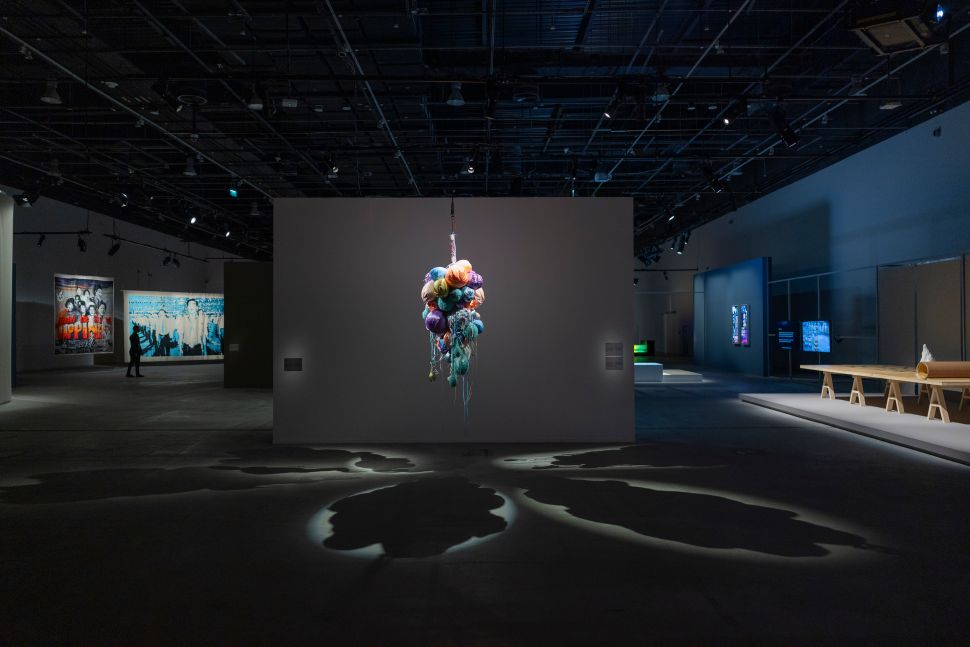 Central hanging sculpture made of colorful tangled textiles and yarns illuminated dramatically against a gray wall, casting soft, organic shadows that spread across the gallery floor.