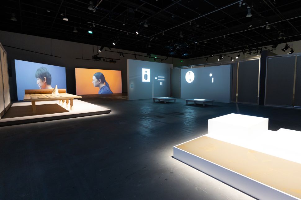 A dimly lit gallery space featuring dual video projections of two individuals in profile—one against a blue background and the other against orange—accompanied by a central wooden table installation. Adjacent walls display minimalistic icon-based digital projections resembling interface elements, while glowing white plinths and low platforms accentuate the futuristic ambiance of the exhibition.
